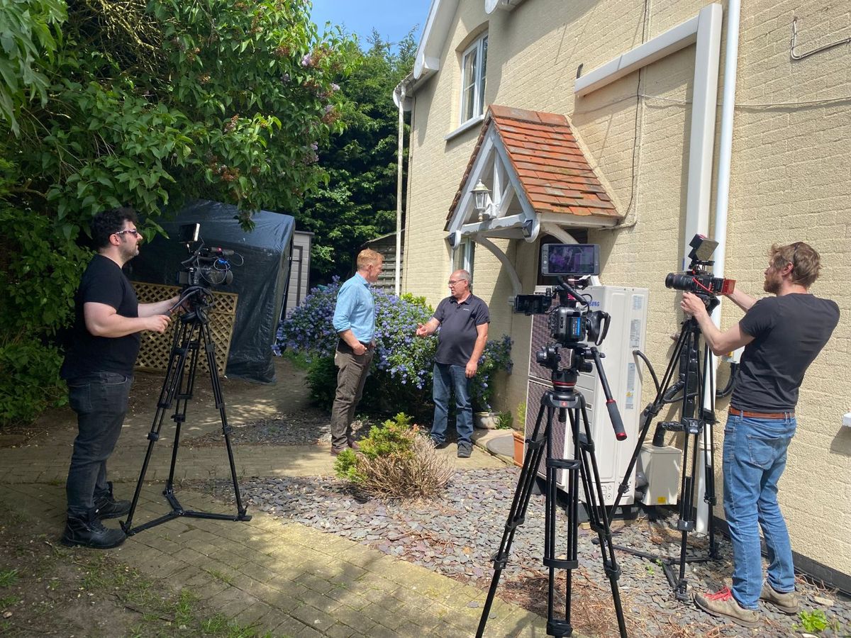 Adam Henson being filmed in conversation beside an air source heat pump, with two camera operators on tripods